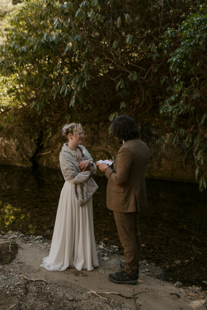 A couple in wedding clothes stands by a river in the woods while the husband reads his wedding vows during their intimate sunrise wedding.