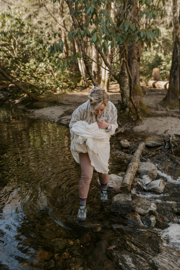 A woman in hiking boots and a wedding dress walks across a creek in the woods.