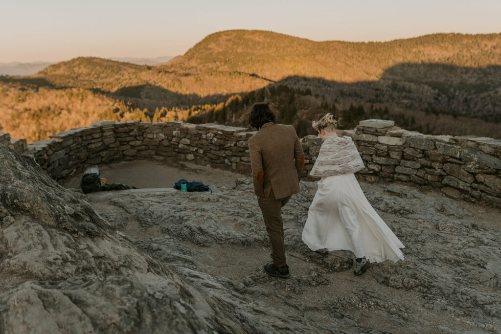 Couple holds hands and walks toward an overlook in the mountains where they’ll have their intimate sunrise wedding.