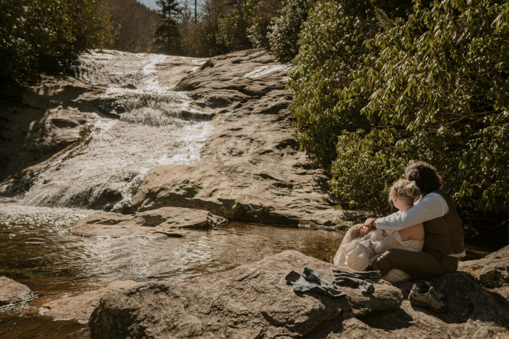 A newlywed couple cuddles on a rock overlooking a waterfall in the forest.