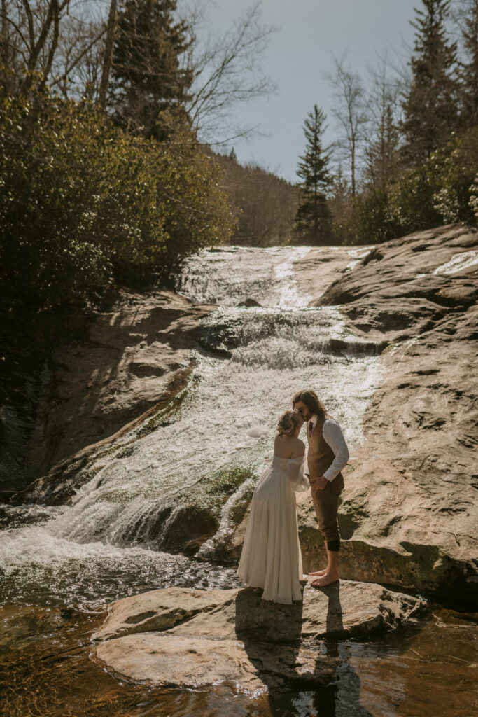 A husband and wife stand on a rock next to a waterfall and kiss in the woods.