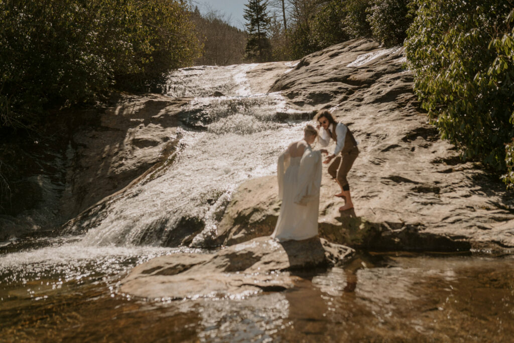 A man helps a woman in a wedding dress walk across a waterfall rock.