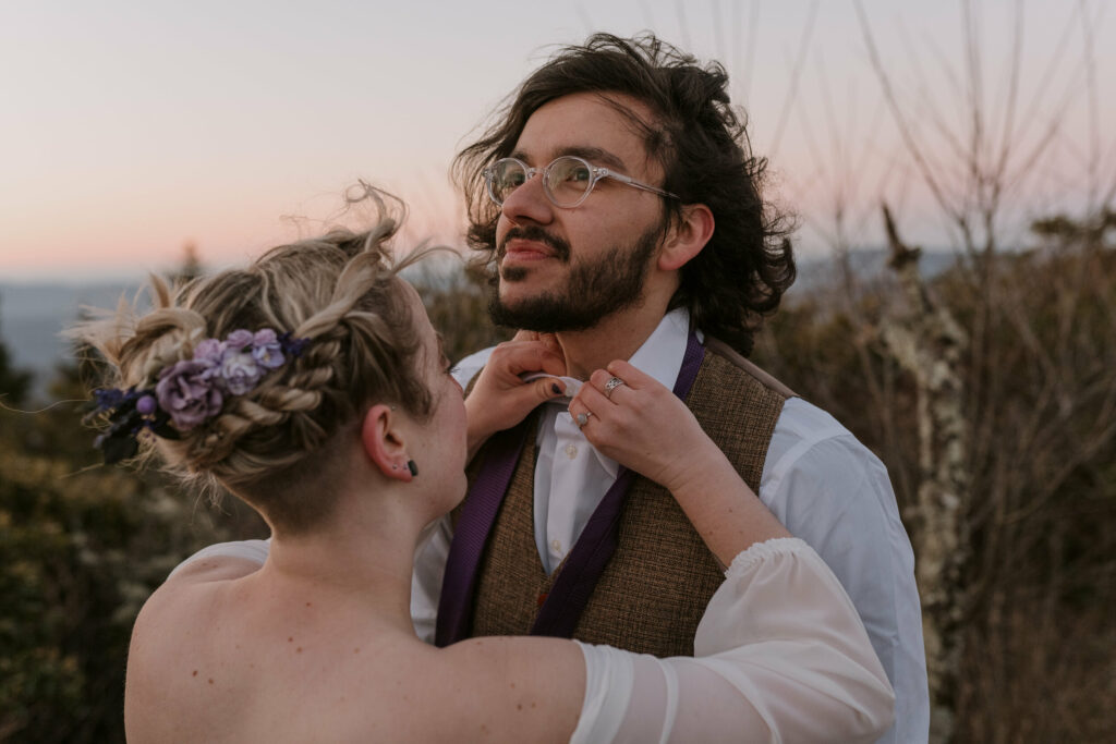 Woman helps her husband with his wedding clothes on top of a mountain at their intimate sunrise wedding.