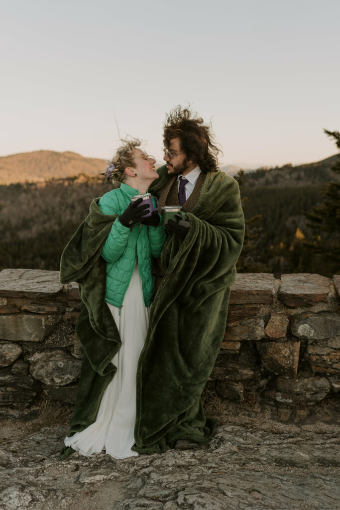 A couple covered in a green velvet blanket and holding mugs of coffee smiles at each other while standing on an overlook in the mountains during their intimate sunrise wedding.