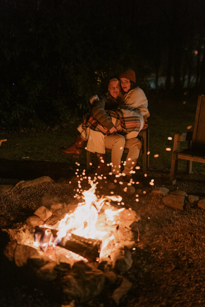 A person holds a woman covered in a plaid blanket while sitting next to a fire in the dark.