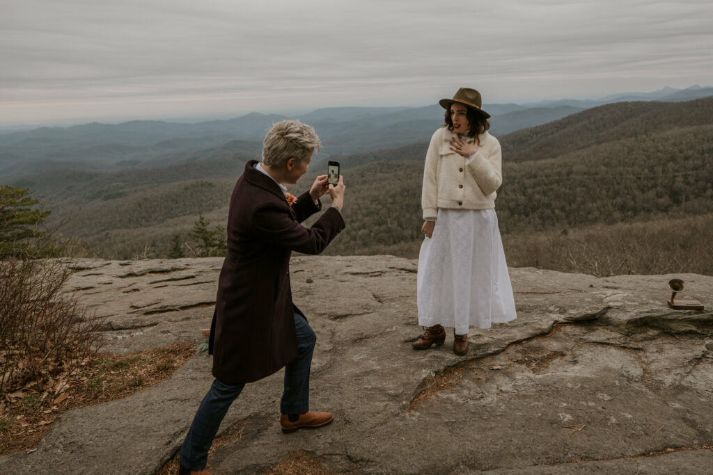 A person takes a cell phone picture of their newly married partner on top of a mountain.