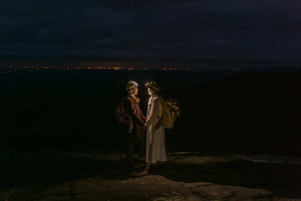 A couple stands on top of a mountain in the dark wearing headlamps in Asheville as the sun sets behind them and you can see the city lights.