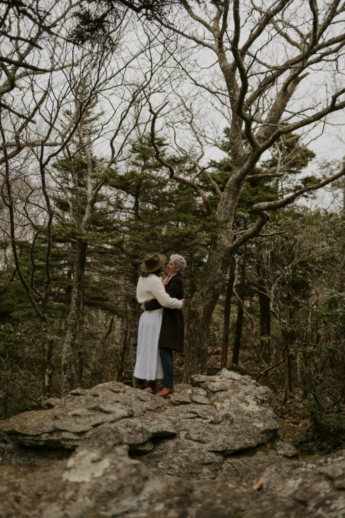 A portrait of a couple embracing on a rock with trees in the background after their winter elopement in the NC mountains.