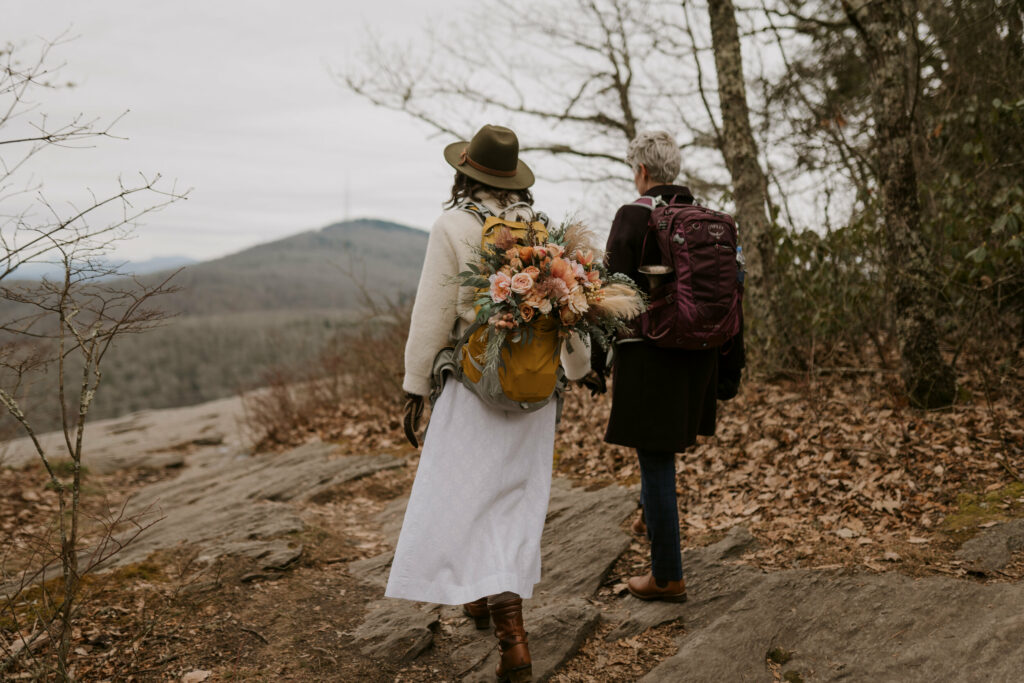 A couple wearing backpacks and wedding clothes walks across a large rock to their elopement ceremony site with mountains in the distance.