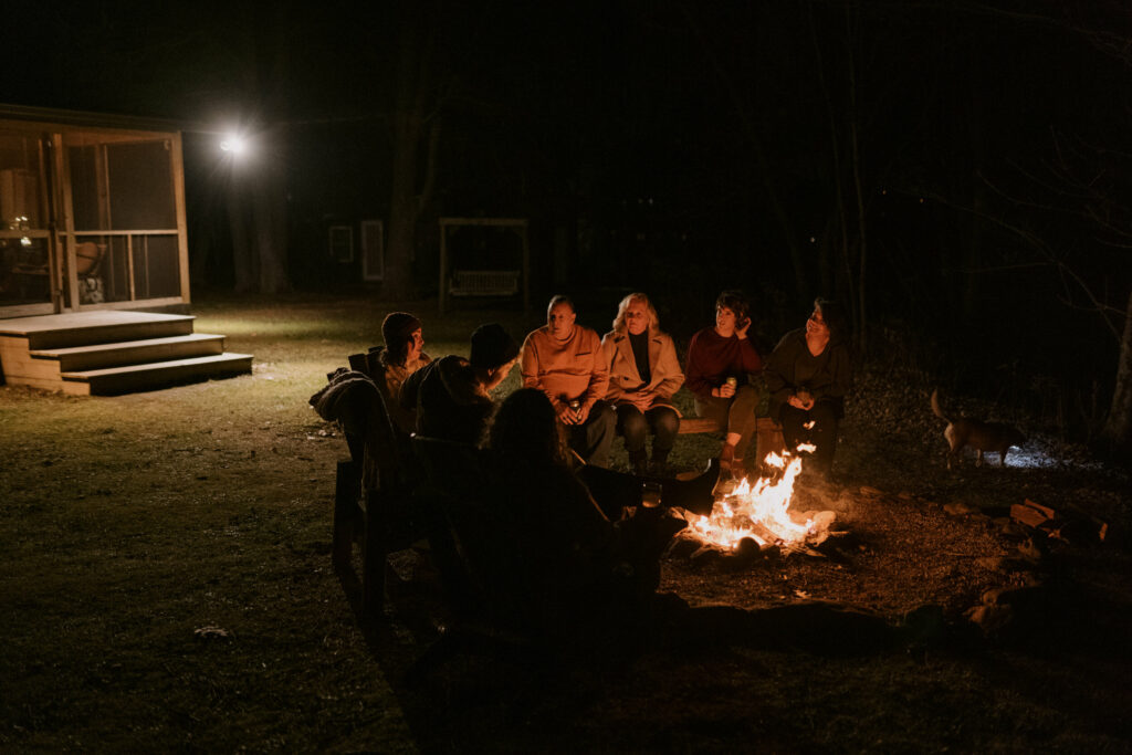 Friends and family sitting around a campfire in the dark with a cabin in the background.