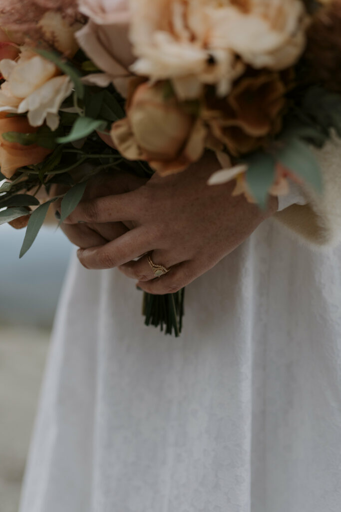 A closeup photograph of a woman’s wedding ring on her hand while holding her bouquet.