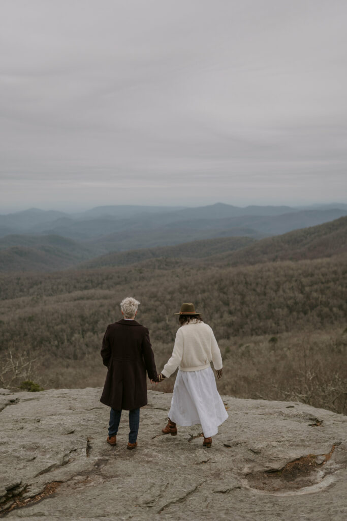 A couple holding hands while standing on top of a rock and overlooking the mountains in wedding attire.