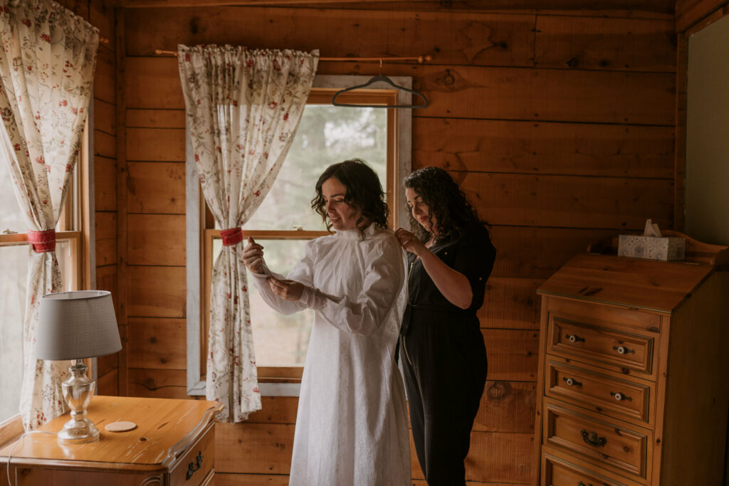 A friend helping a woman put on her wedding dress before her elopement day inside a cabin.