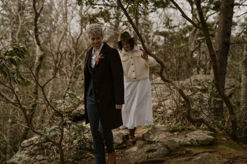 A couple hikes through bushes on their way up a mountain wearing wedding clothes.