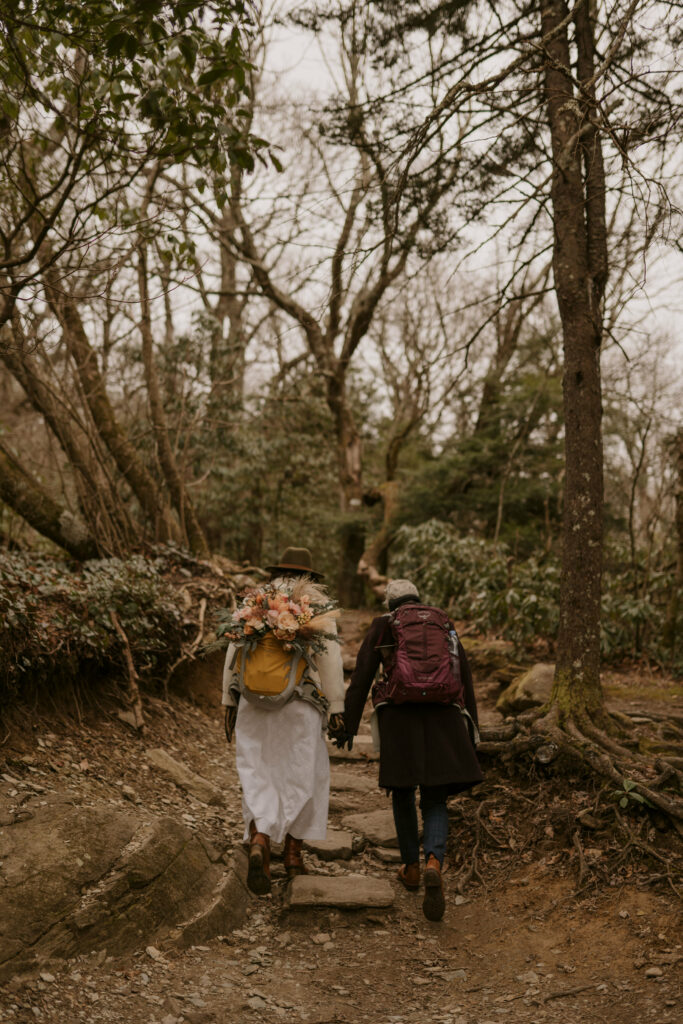 A couple holds hands and walks up a forested mountain hiking path while wearing backpacks.