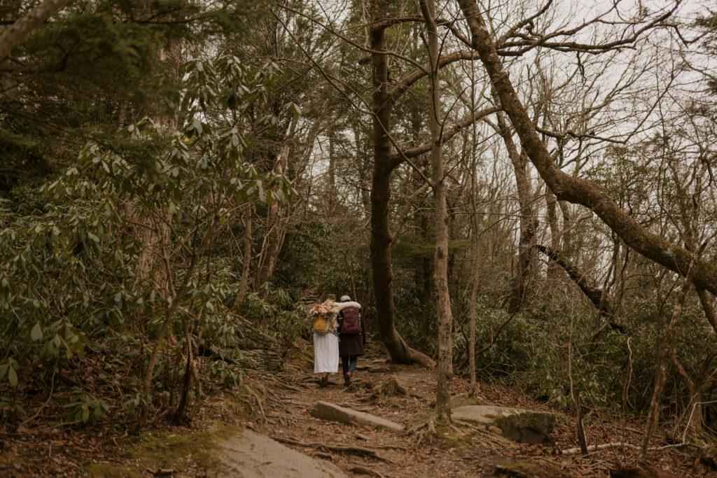 A couple wearing backpacks hikes up to elopement destination in NC mountains surrounded by trees.