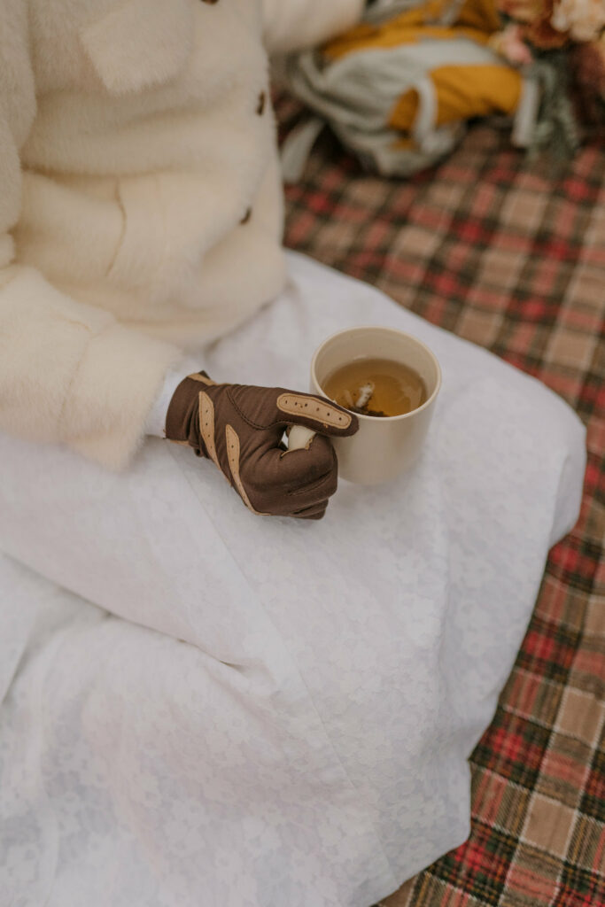 A woman wearing gloves holds a mug full of tea while sitting on a plaid blanket and wearing a wedding dress.