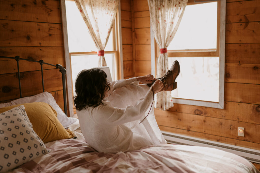A woman laces up boots while wearing a wedding gown inside a cabin.