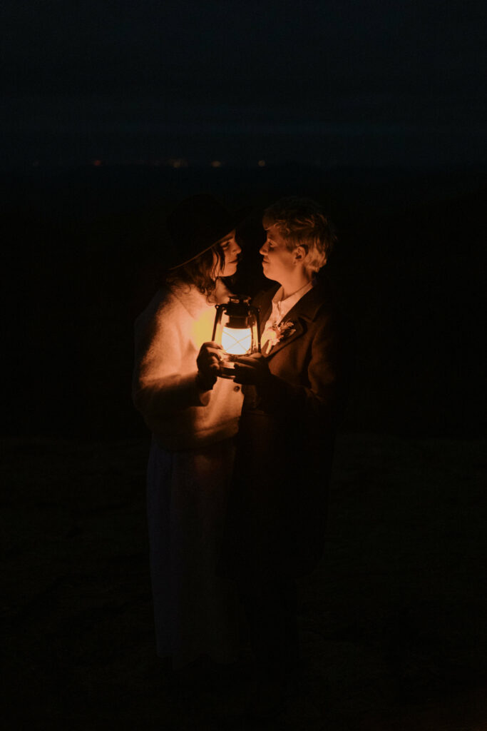 A couple sits in the dark and looks at each other while illuminated by a lantern.
