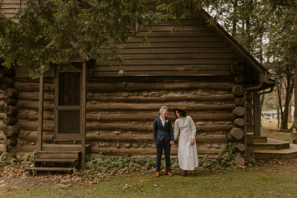 A laughing couple standing outside a log cabin while holding hands.