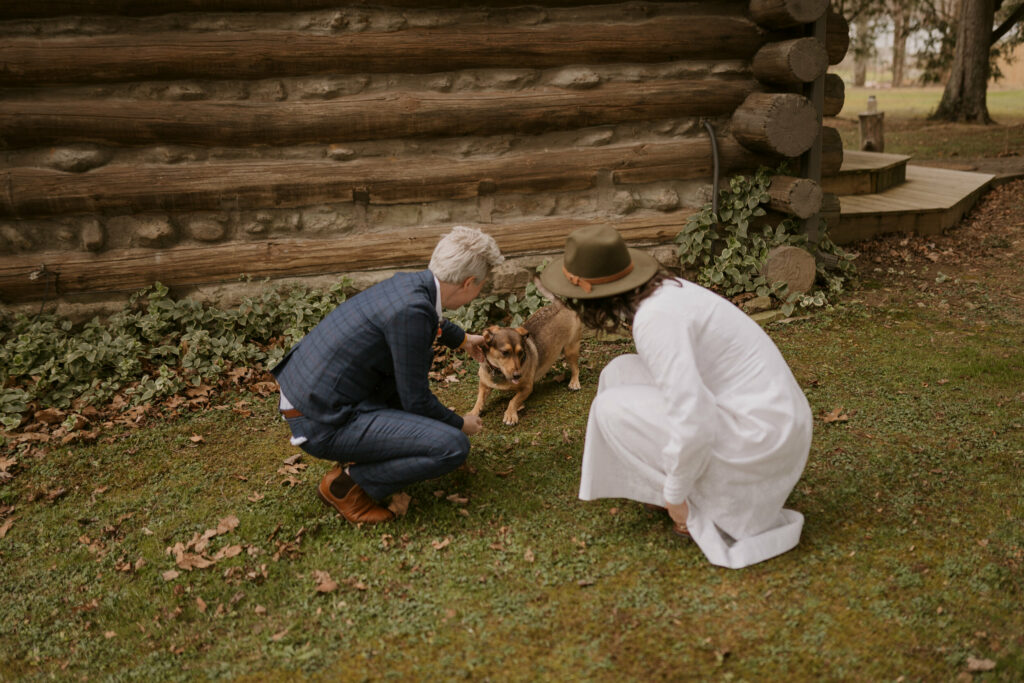 A couple talks to their little dog outside a log cabin.