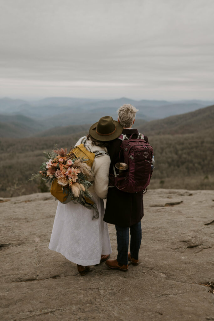 A woman in a dress leans her head on a person in a suit while they both stand on a rock and overlook a mountain vista.
