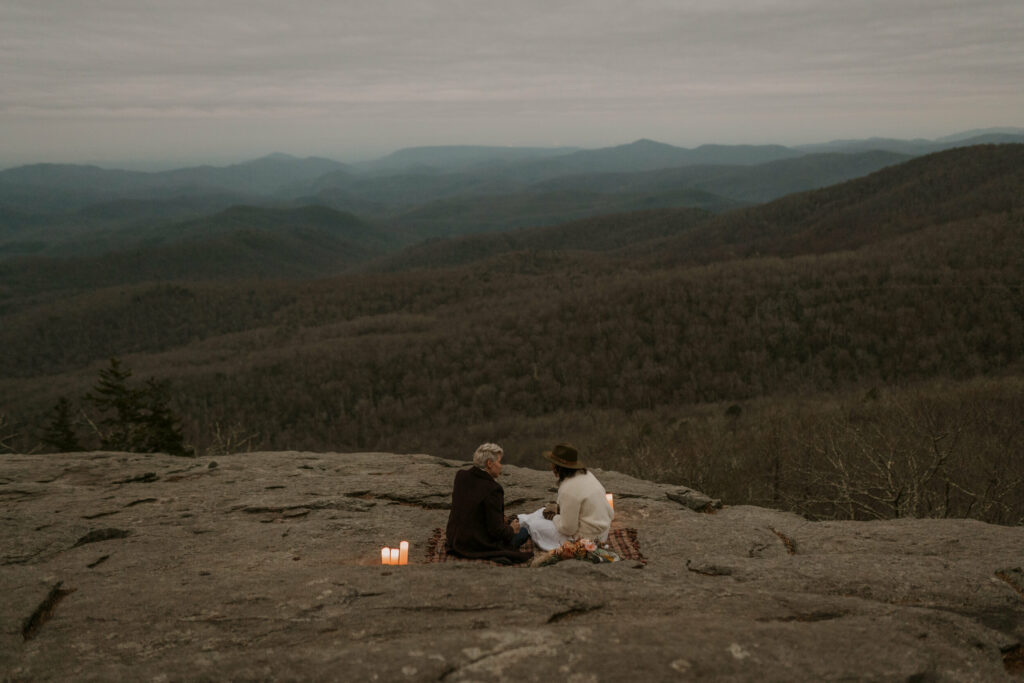A couple sits overlooking the mountains on top of a blanket with candles.