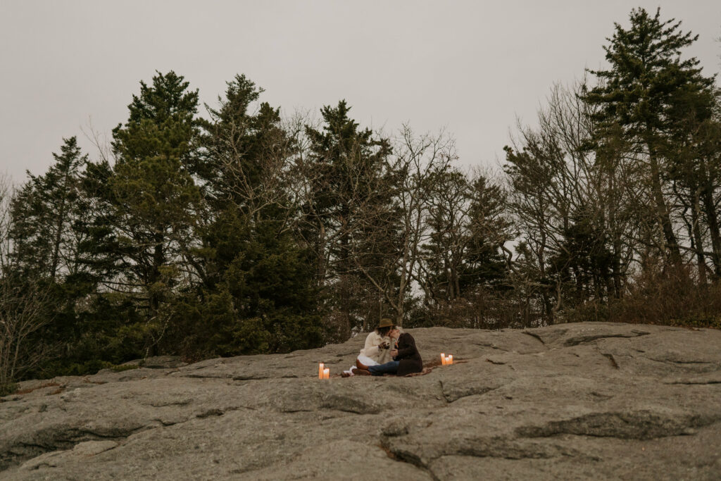 A couple sits on top of a rock with pine trees in the background while they drink tea on a picnic blanket.