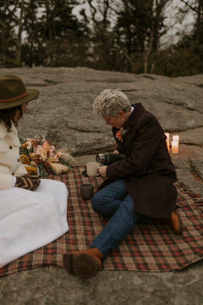 A person pours tea out of a mug on top of a plaid blanket in the winter on a mountaintop.
