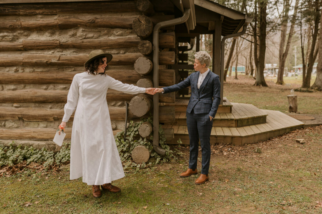 A couple reaches for each other and looks at each other after first look outside a cabin.