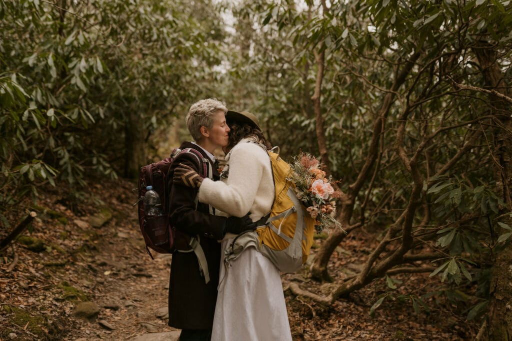 A couple wearing backpacks hugs among the rhododendrons while hiking up to a mountain in Asheville.