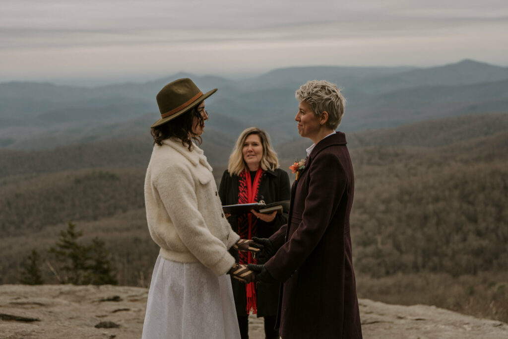 A couple saying their vows during their winter elopement on top of a mountain with an officiant behind them and a mountain range surrounding them.