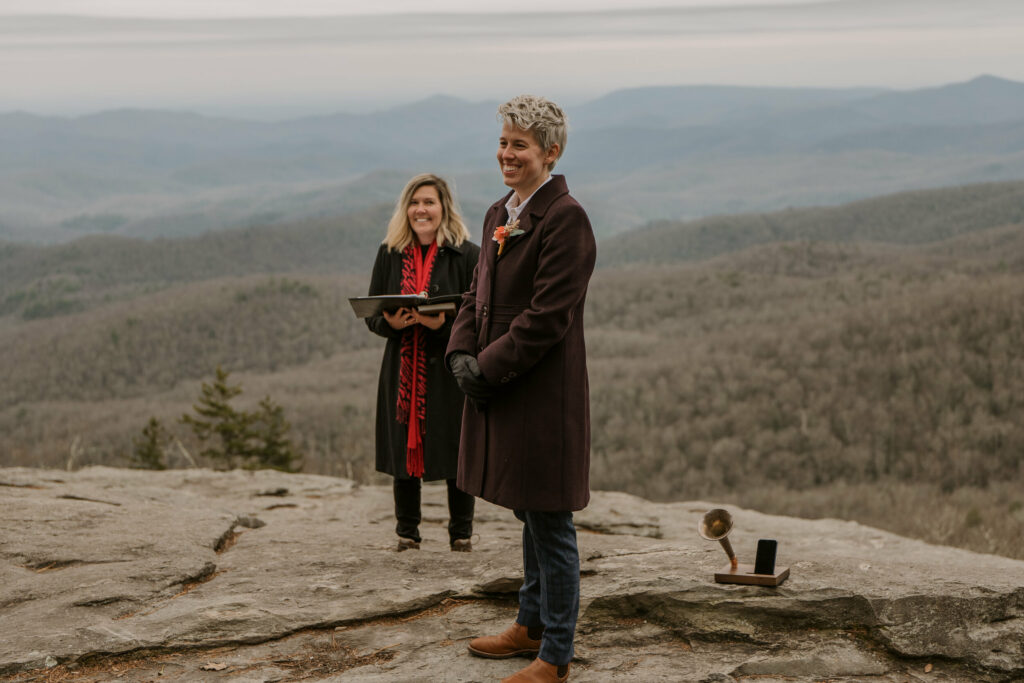 A person in a suit and coat stands smiling on a mountaintop with a wedding officiant in the background.
