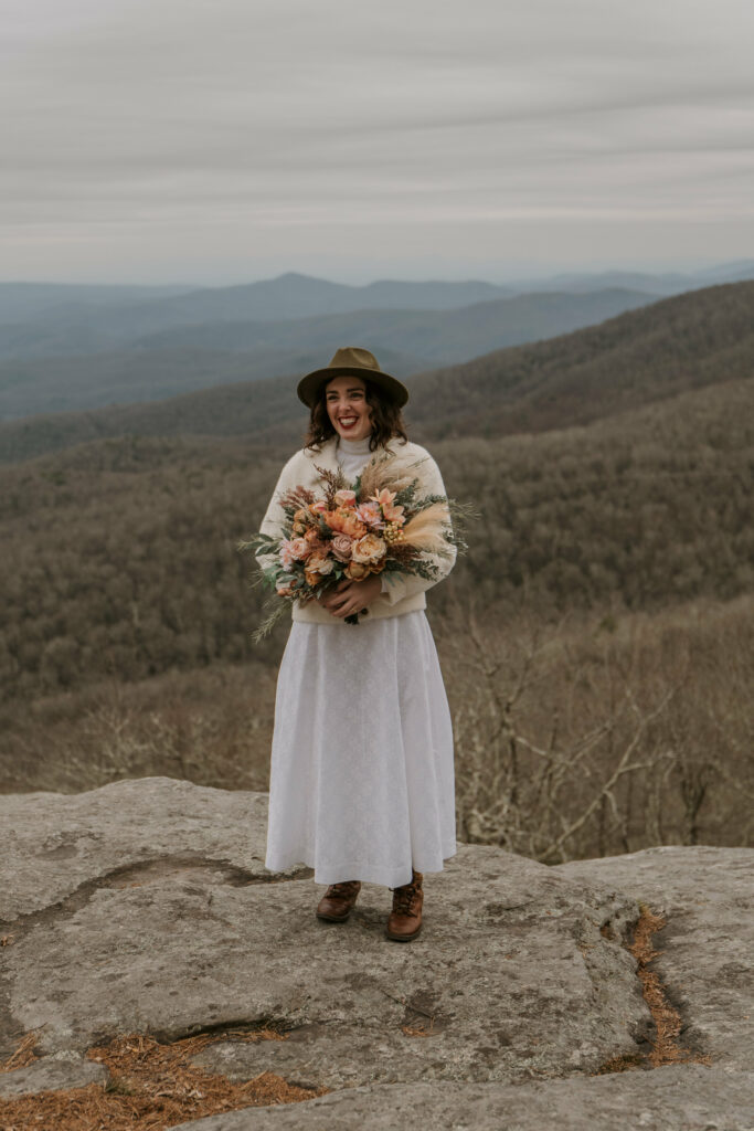 A woman smiling while holding a bouquet of flowers and wearing a white dress and brown hat surrounded by mountains.