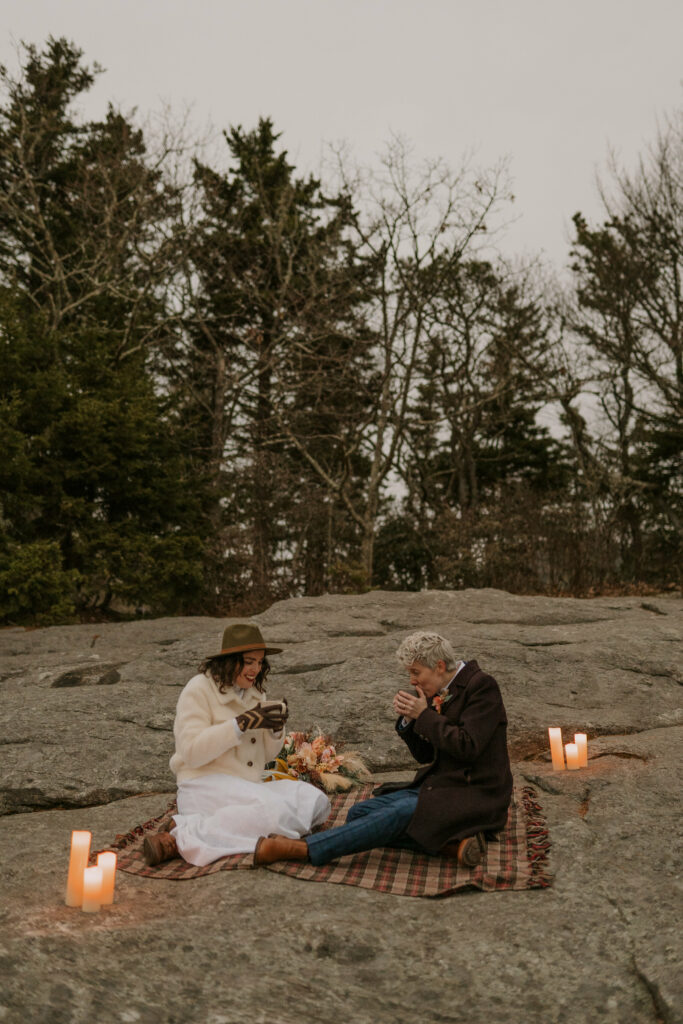 A couple drinks tea on top of a mountain in the winter on a plaid blanket surrounded by candles.