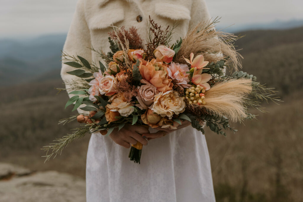 A close up of a bride holding a faux floral bouquet that is orange and pink and green with mountains in the background during a winter blue ridge parkway wedding.