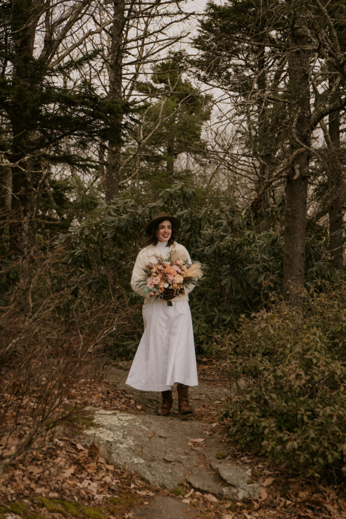 A woman in a wedding dress and hat carrying a bouquet walks down a mountain path toward her elopement ceremony.