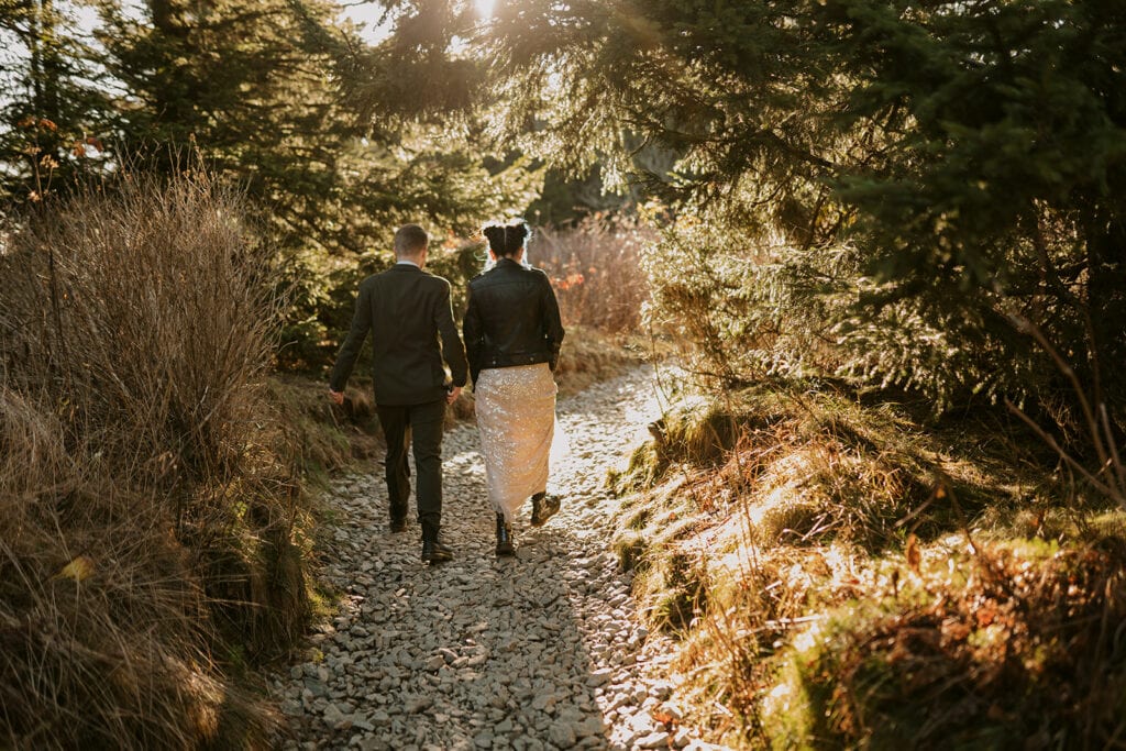A couple is walking through a gravel path in the forest with wedding clothes on and the sun shining on them.