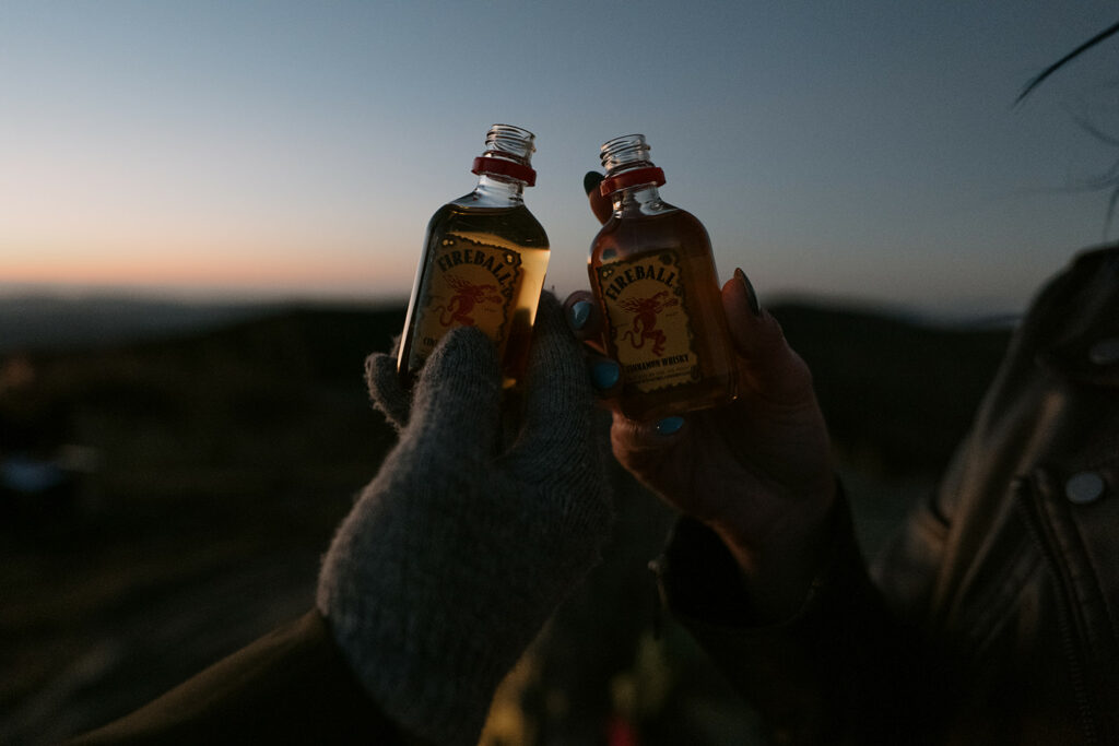 Two hands are holding tiny fireball shot bottles close together outdoors in blue hour light.