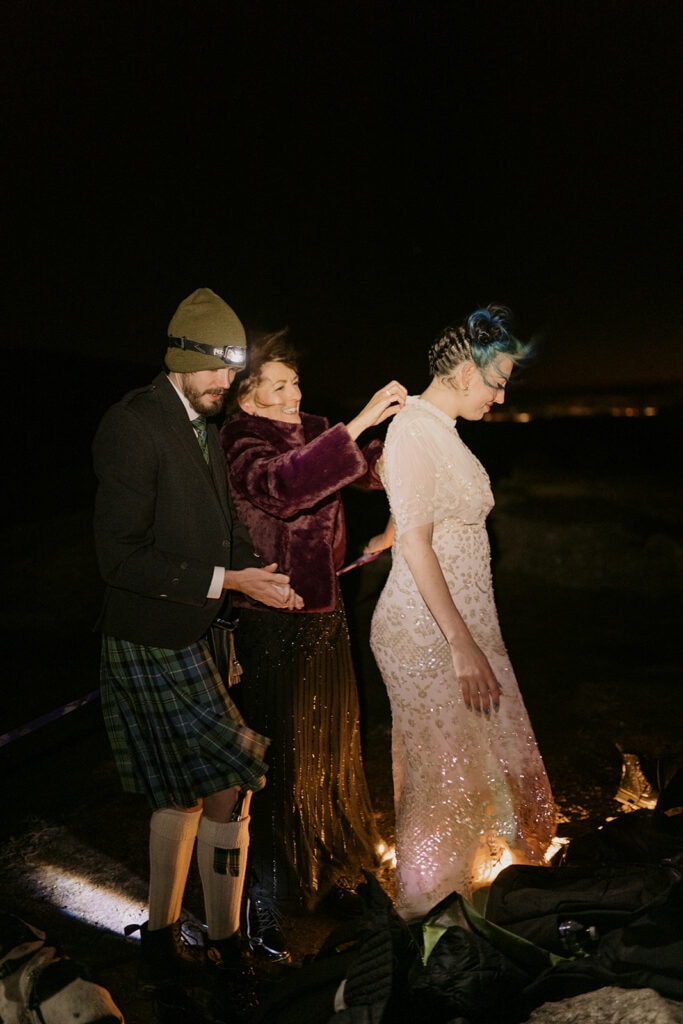 A bride is getting dressed in a sparkly wedding dress at night with the help of her mother and brother.
