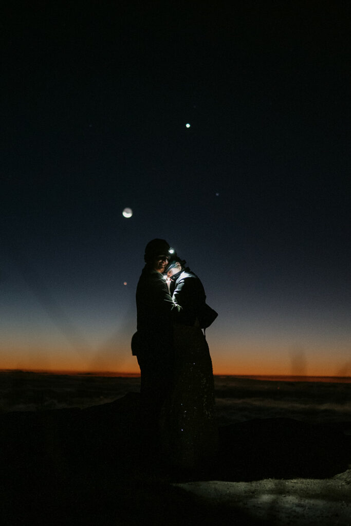 A couple snuggles together with headlamps on at Black Balsam Knob for their wedding with the sun about to rise and the moon and stars above them.