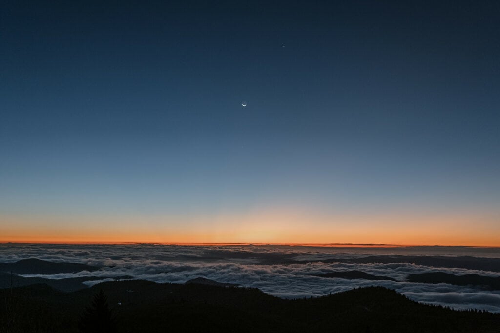 A landscape of mountains, a cloud inversion, the moon and stars right before the sun rises. The sky is blue and orange.