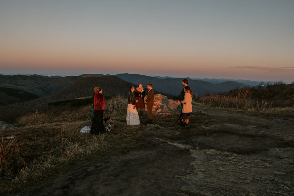 A small group of people are gathered together on a mountaintop at sunrise on Black Balsam Knob for a small wedding celebration. There are 7 people and 2 dogs.