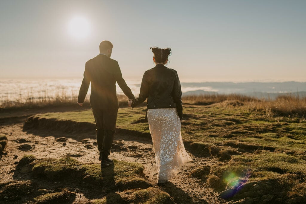 A couple is holding hands and walking across a mountaintop at Black Balsam Knob with the sunshine bright behind them. One person is wearing a suit and the other is wearing a wedding dress and leather jacket.
