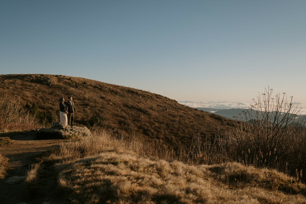 A couples is standing on a rock at Black Balsam Knob holding hands and looking out over the landscape in their wedding clothes.