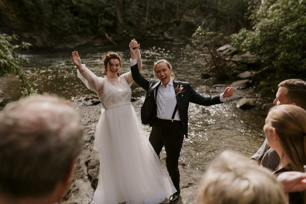 A couple is holding hands in celebration at the end of their elopement with big smiles on. One female is wearing a suit and the other is wearing a white dress.