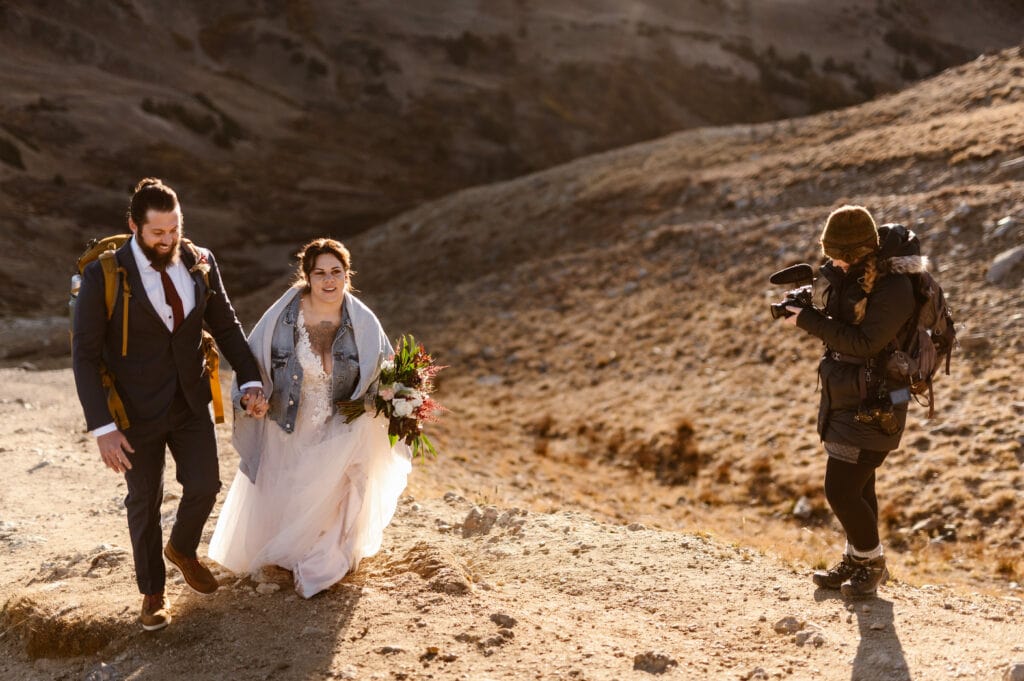 An elopement videographer is standing on a trail filming a couple hiking up a mountain together holding hands and laughing.