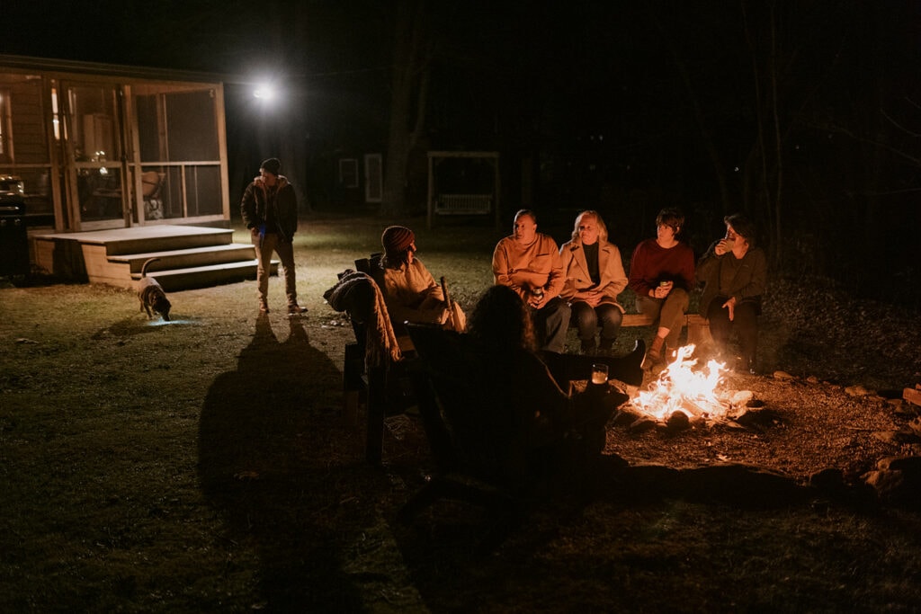 A group of people sit around a campfire chatting in the backyard of a house at night time.