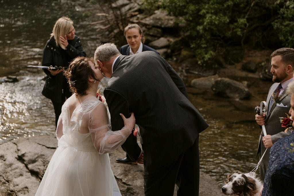 A bride is kissing her family member (father) on the cheek as he drops her off at her elopement ceremony spot by a creek. Others are standing around watching