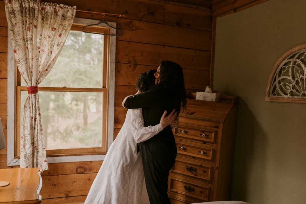 Two family members, sisters are hugging in a cabin style room. One is wearing her elopement dress and the other is in a black outfit.